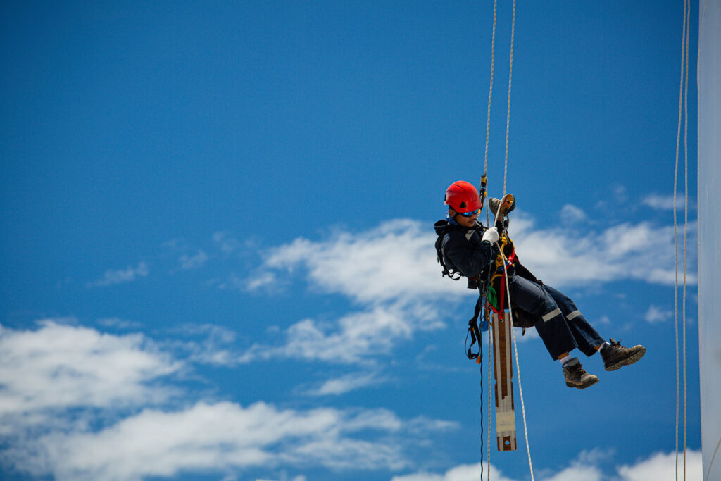 Worker in safety gear suspended by ropes against blue sky.